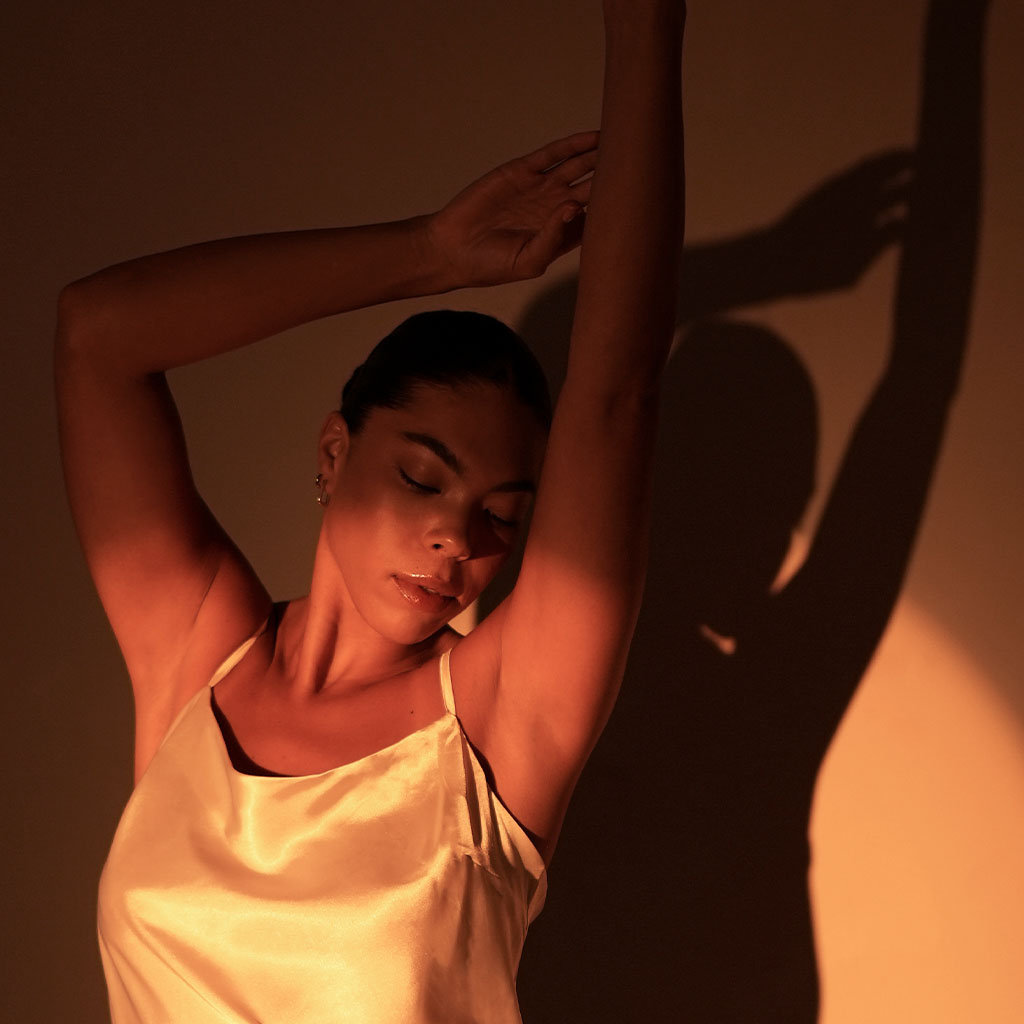 Eine Frau in einem glänzenden, hellen Satin-Top posiert mit erhobenen Armen. Ihr Gesicht ist entspannt, die Augen geschlossen, während warmes kupferfarbenes Licht Schatten an der Wand hinter ihr wirft.