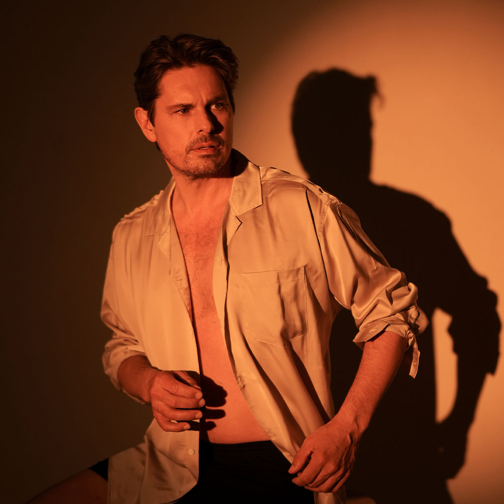 Ein Mann mit ernstem Blick trägt ein offenes beiges Hemd über nacktem Oberkörper. Er posiert im warmen, kupferfarbenen Licht, wodurch sein Schatten deutlich an der Wand hinter ihm zu sehen ist.