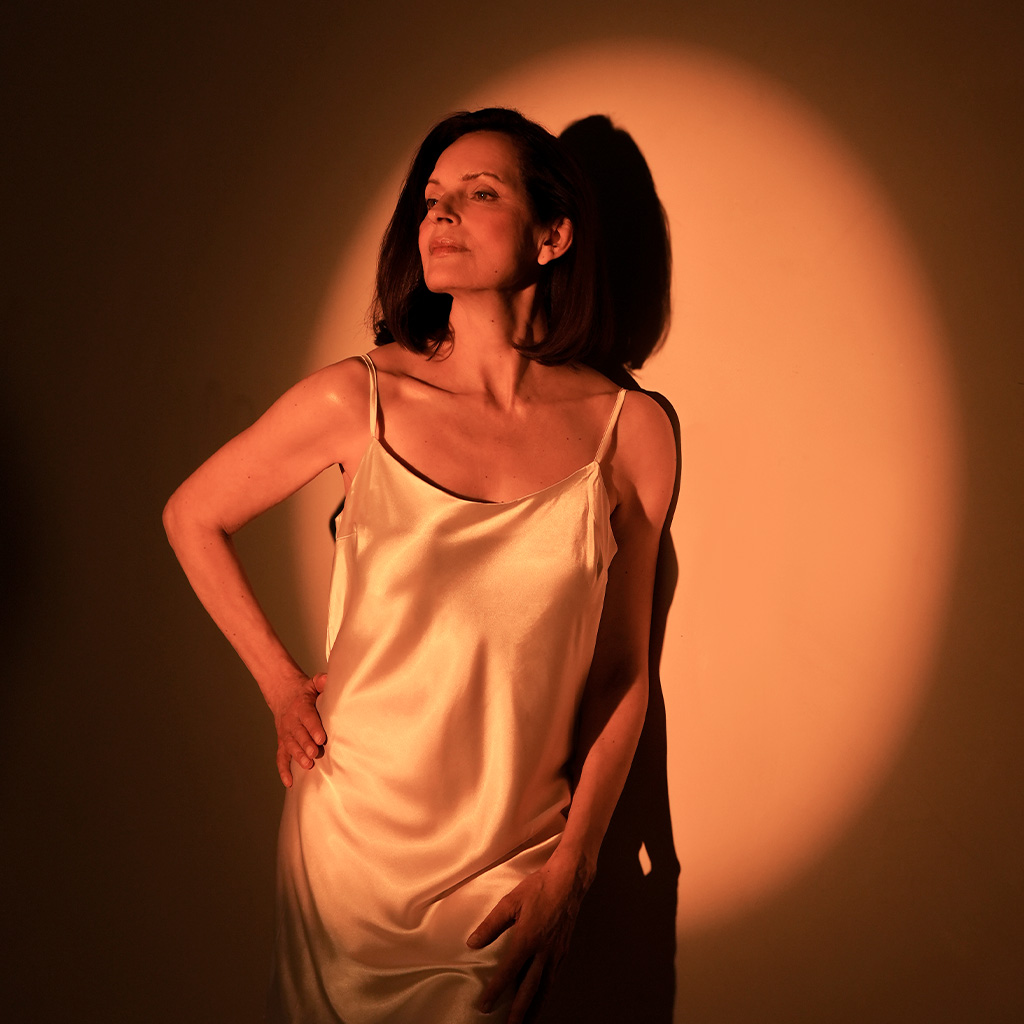 Eine Frau mit schulterlangem dunklem Haar trägt ein seidiges, helles Kleid. Sie steht selbstbewusst im kupferfarbenen Lichtkegel vor einer Wand, mit einer Hand in der Hüfte und dem Blick nach oben gerichtet.