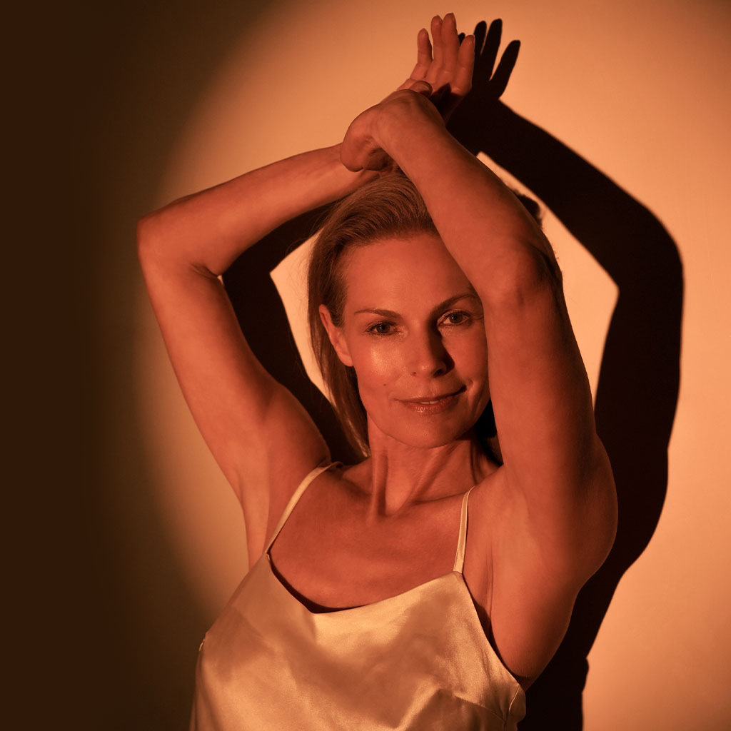Eine Frau mit blonden Haaren trägt ein seidiges Trägertop und hebt ihre Arme elegant über den Kopf. Warmes, kupferfarbenes Licht beleuchtet sie, während ihr Schatten deutlich an der Wand hinter ihr erscheint.